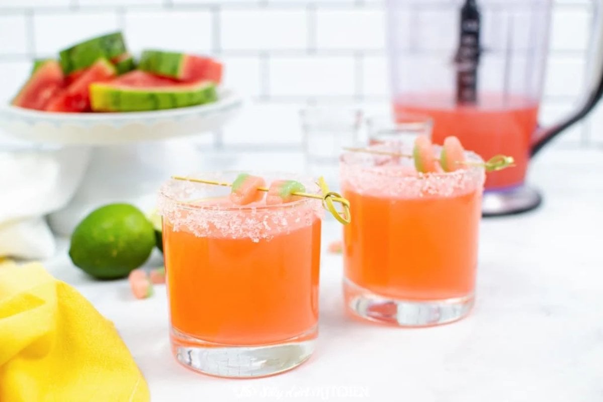 Two glasses of orange-pink beverages with salted rims and candy garnishes are on a countertop. In the background, there is a plate of watermelon slices and a blender, hinting at refreshing watermelon recipes. A lime and a yellow cloth are nearby.