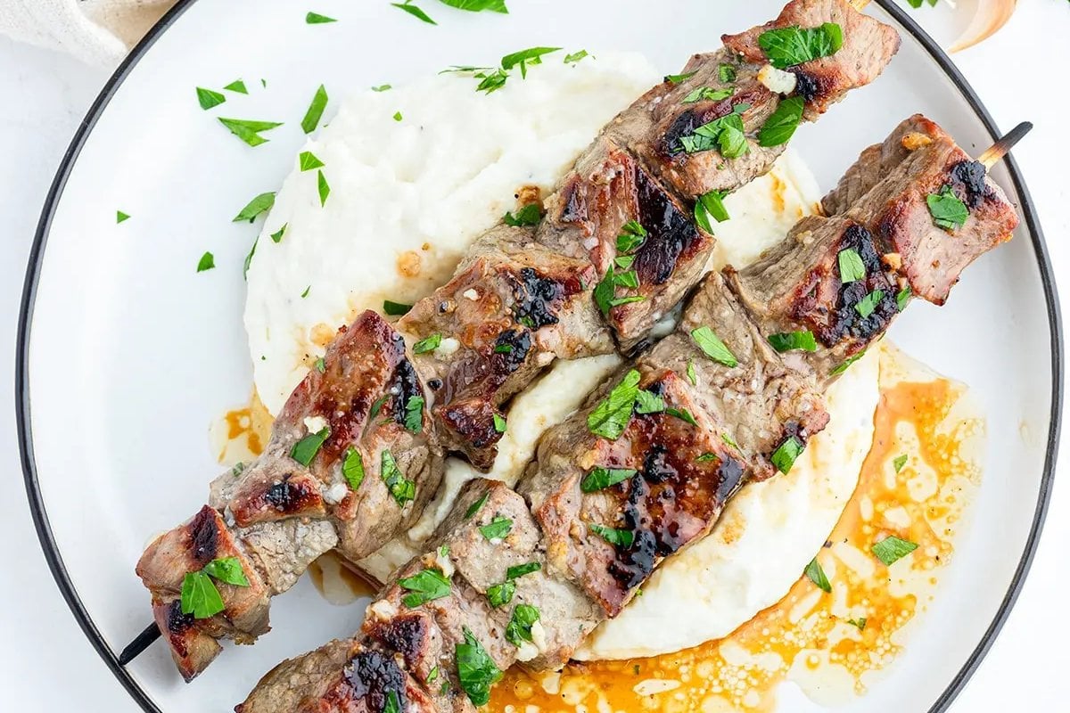 Two grilled meat skewers garnished with herbs lie on a bed of creamy mashed potatoes, reminiscent of classic steak recipes, presented on a white plate.