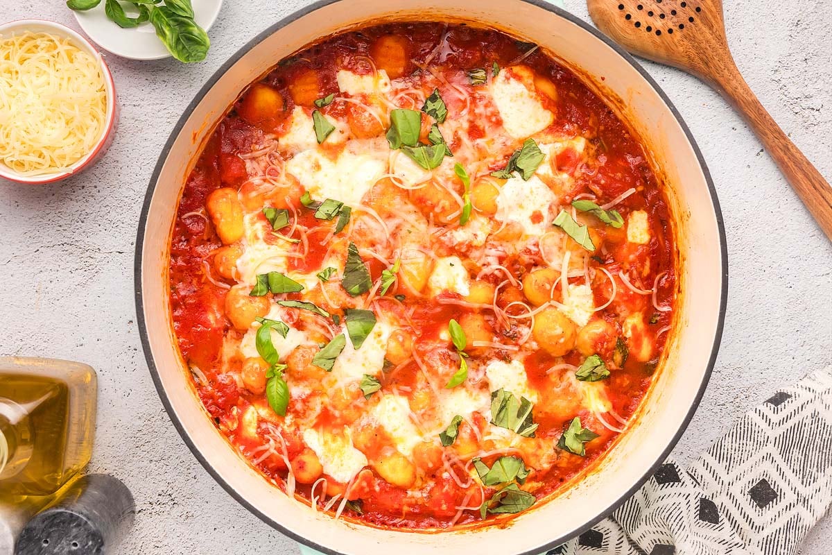 A pot of gnocchi with tomato sauce is garnished with melted cheese, fresh basil, and shredded Parmesan. The meal is accompanied by a wooden spoon, a small bowl of grated cheese, a bottle of olive oil, and a kitchen towel, all set on a light-colored surface.