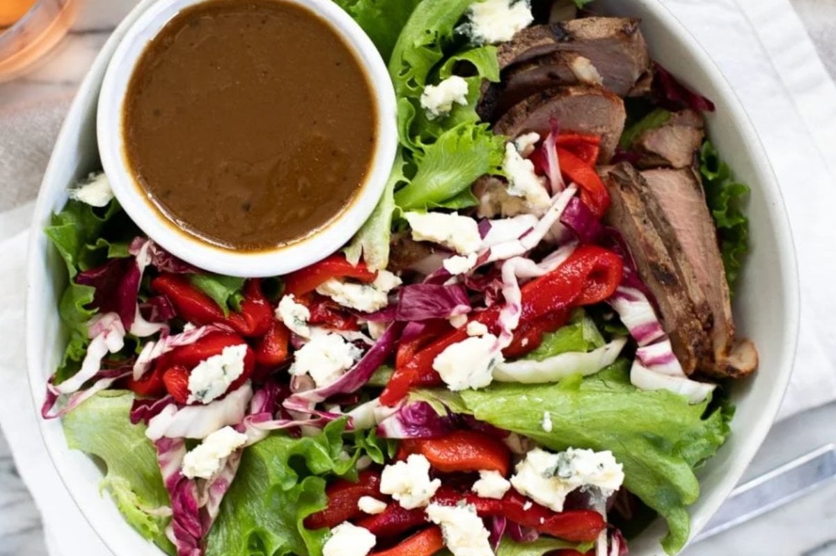 A bowl of salad featuring mixed greens, slices of grilled steak, red bell peppers, crumbled cheese, and a small bowl of brown dressing. Perfect for those who love hearty steak recipes with a fresh twist.