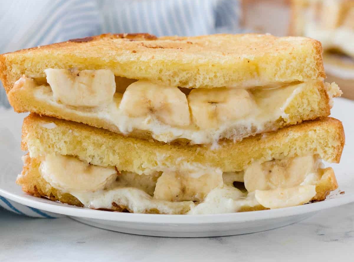 Grilled sandwich halves filled with melted cheese and banana slices are stacked on a white plate, offering a unique twist on classic grilled cheese recipes.