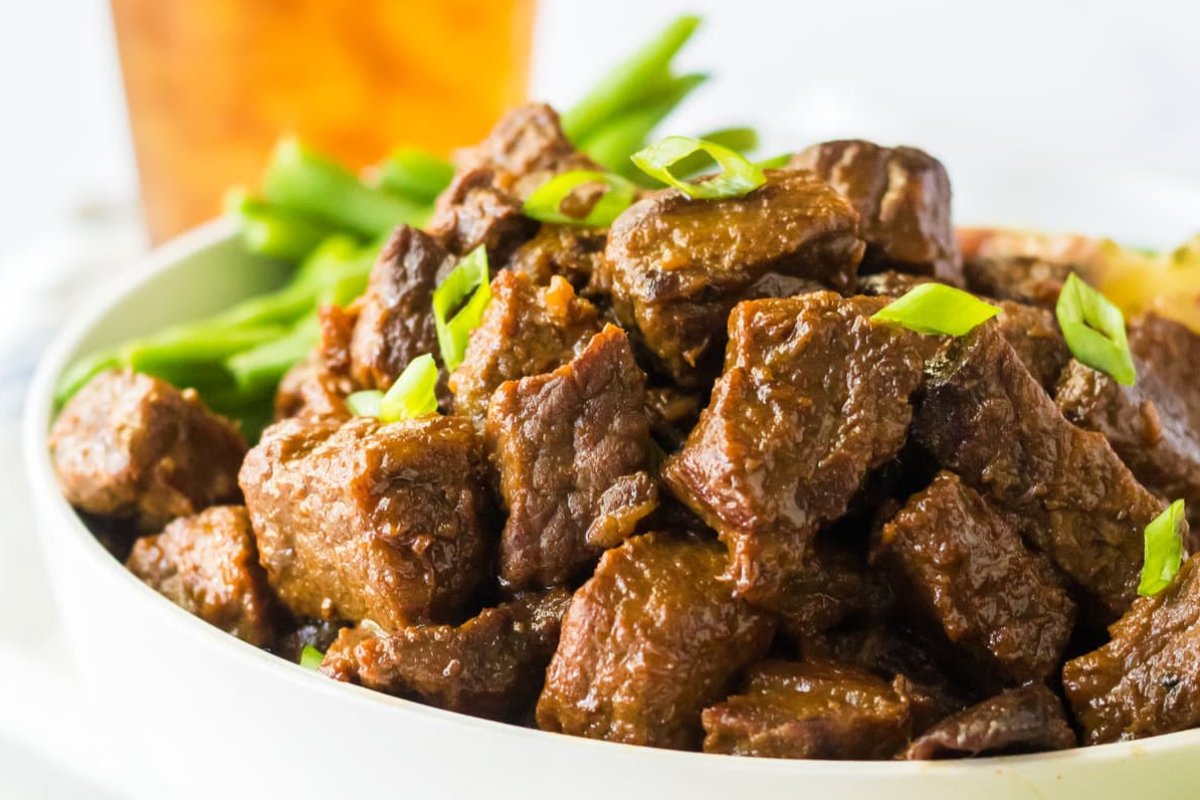 A bowl filled with cooked beef chunks garnished with chopped green onions, reminiscent of the best steak recipes, with green beans in the background.