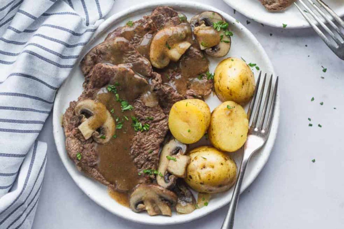 A plate featuring sliced steak in mushroom sauce, accompanied by whole boiled potatoes and garnished with chopped herbs. Two forks and a striped napkin are visible beside the plate, making it a picture-perfect example for any steak recipes collection.