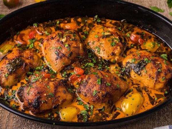 A black baking dish filled with creamy lemon chicken thighs with spinach, garnished with herbs and surrounded by a colorful array of vegetables in a seasoned broth. A cloth napkin and side dishes are partially visible.