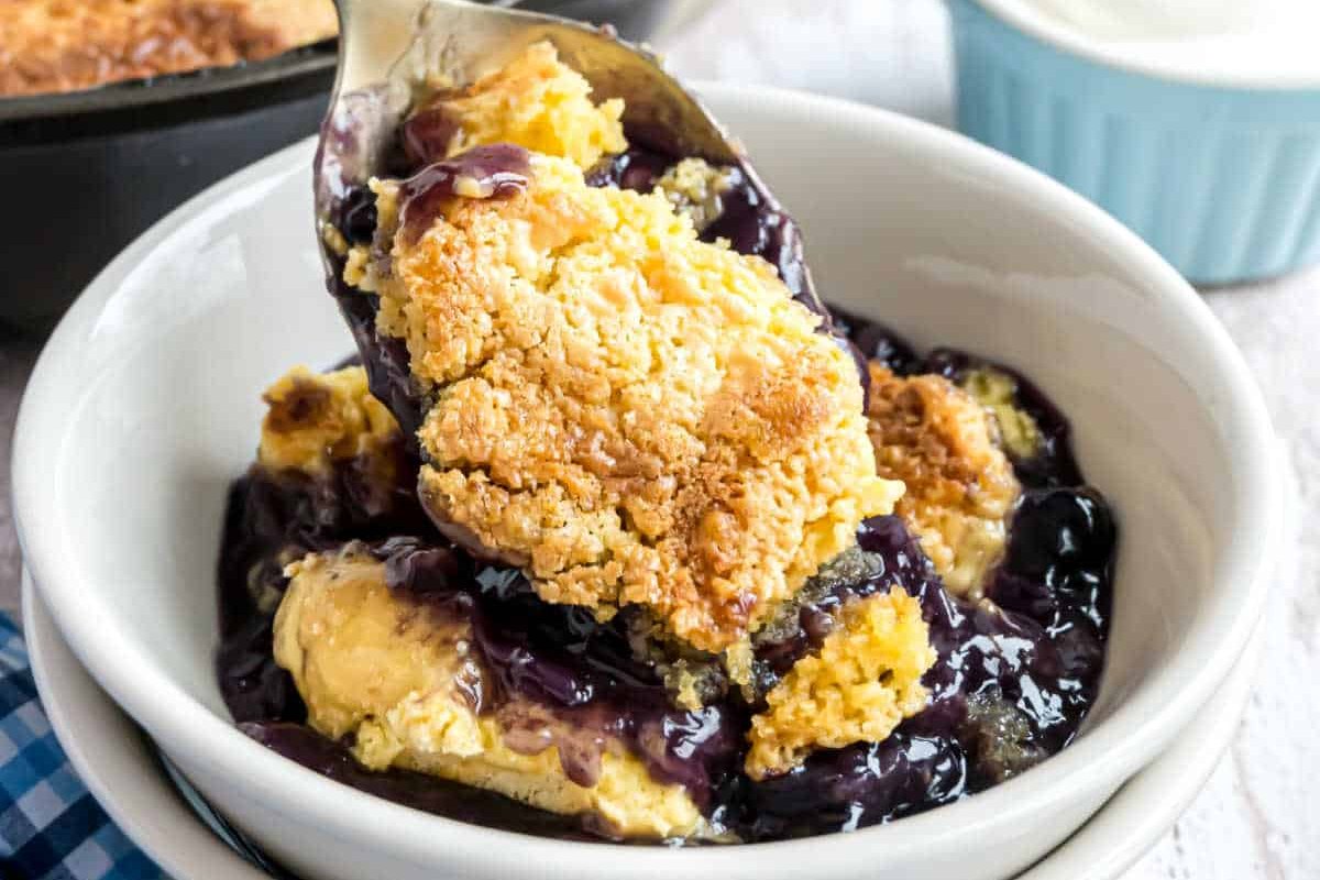 A close-up of a spoon lifting a portion of blackberry cobbler with a golden-brown crust from a white bowl, perfect for those exploring new blueberry recipes.