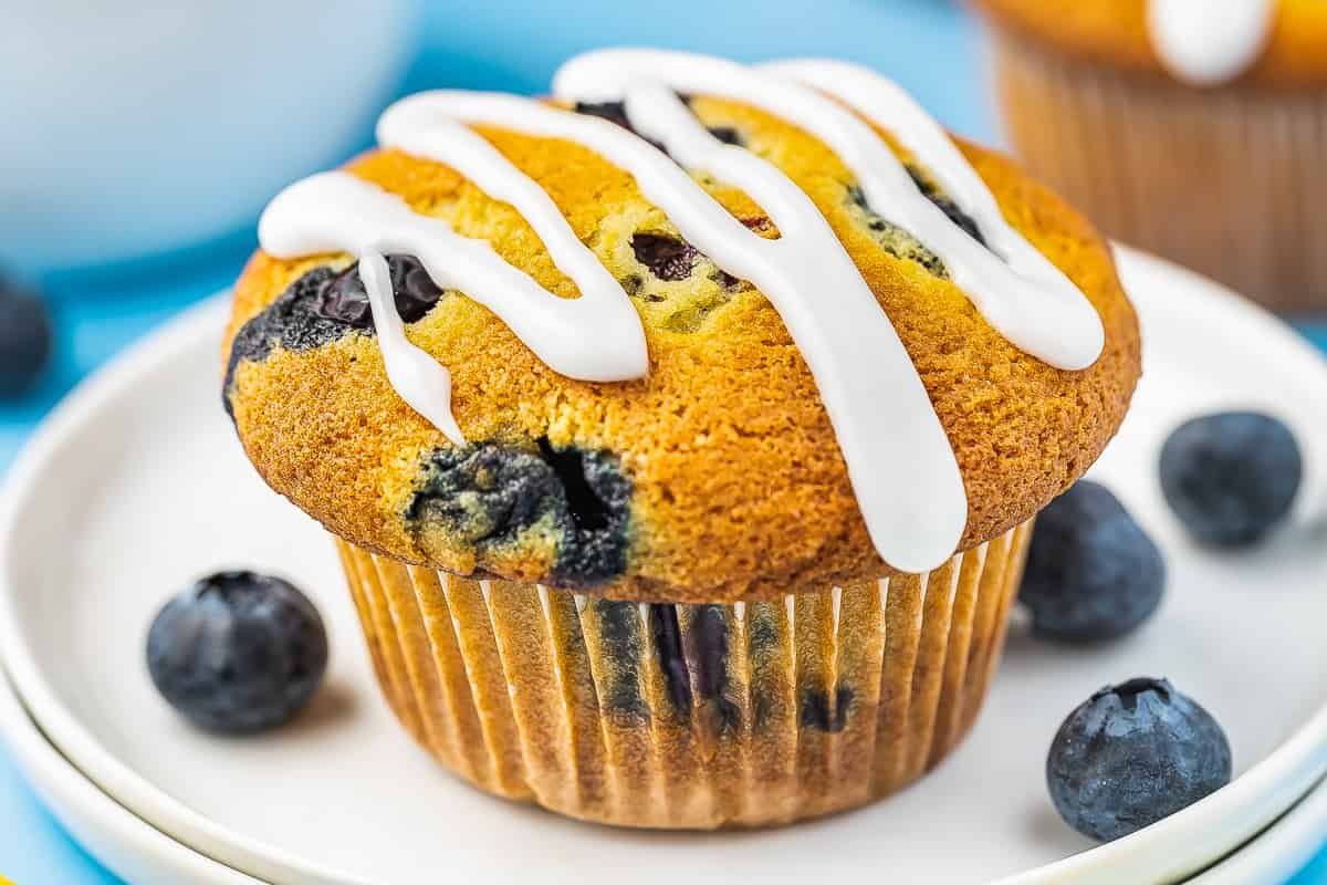 A blueberry muffin with white icing drizzled on top, sitting on a white plate with scattered blueberries around it, offering a delightful twist to traditional lemon desserts.