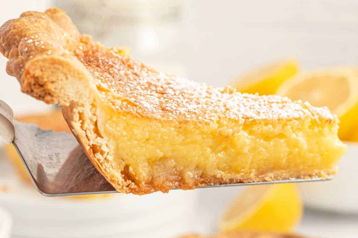 A close-up of a slice of lemon pie with a flaky crust, topped with powdered sugar. Lemons are visible in the background, providing a refreshing touch to this classic lemon dessert.