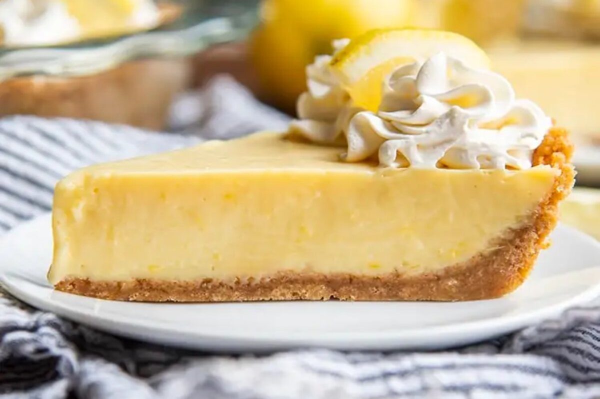 A slice of lemon cream pie with a graham cracker crust, topped with whipped cream and a lemon slice, on a white plate—just one of the many enticing lemon desserts to indulge in.