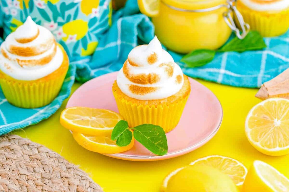 A lemon cupcake with swirled frosting sits on a pink plate, accompanied by lemon slices and a leaf. Other similar lemon desserts, a lemon, and a jar are in the background on a bright yellow and blue setup.