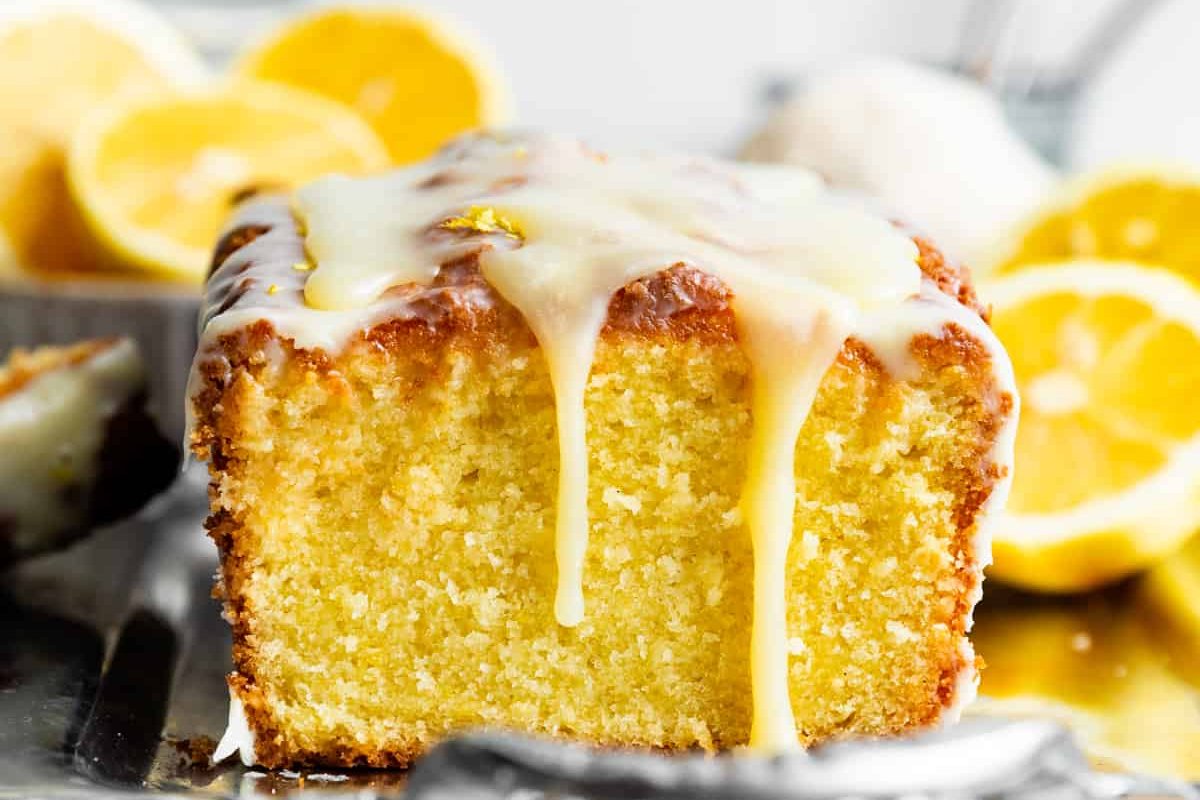 A sliced loaf cake with lemon icing drips from the top, surrounded by halved lemons in the background, making it the perfect addition to your collection of lemon desserts.