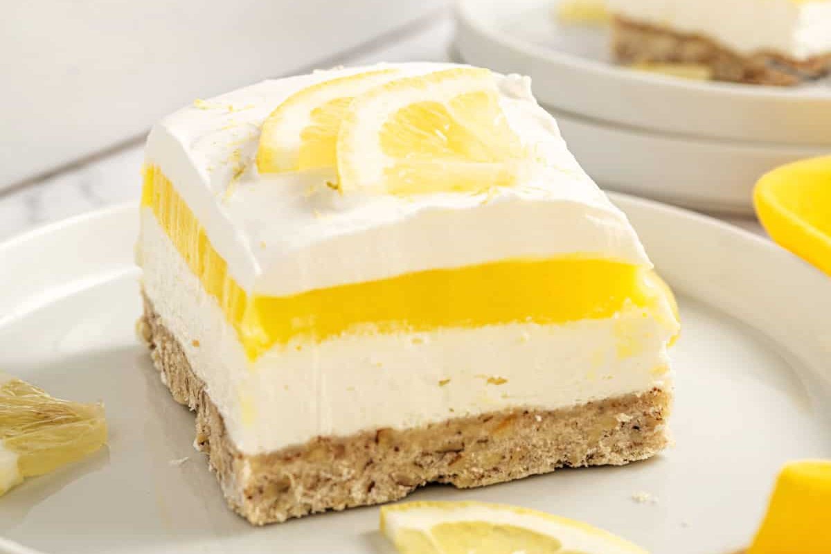 A slice of layered lemon dessert with whipped cream topping and lemon slices, served on a white plate. This delectable creation, one of the finest lemon desserts, features a crumbly crust, a creamy middle layer, and a vibrant yellow lemon layer.