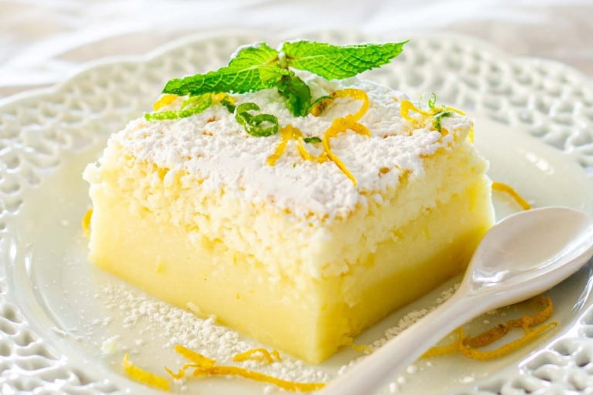 A slice of lemon cake, one of the finest lemon desserts, is topped with powdered sugar, lemon zest, and a sprig of mint on a decorative white plate with a matching white spoon.
