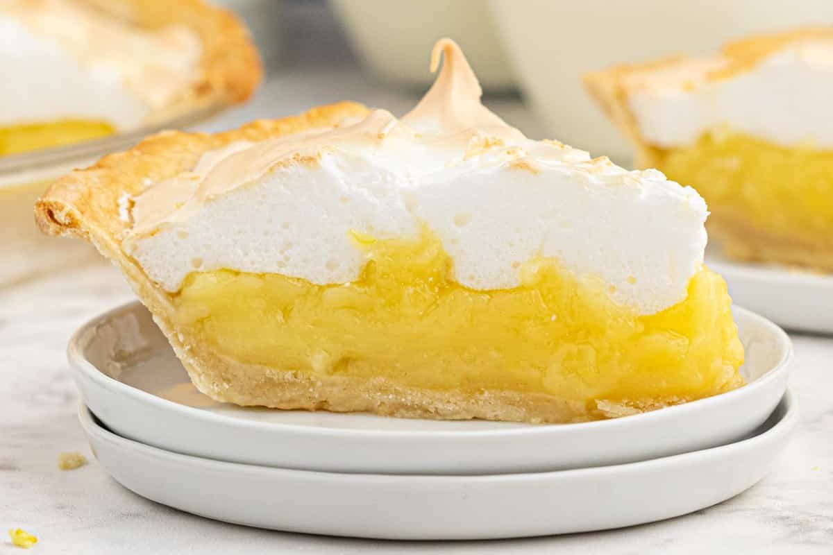 A slice of lemon meringue pie with a light brown crust, yellow lemon filling, and fluffy white meringue topping is the epitome of classic lemon desserts, served perfectly on a white plate.