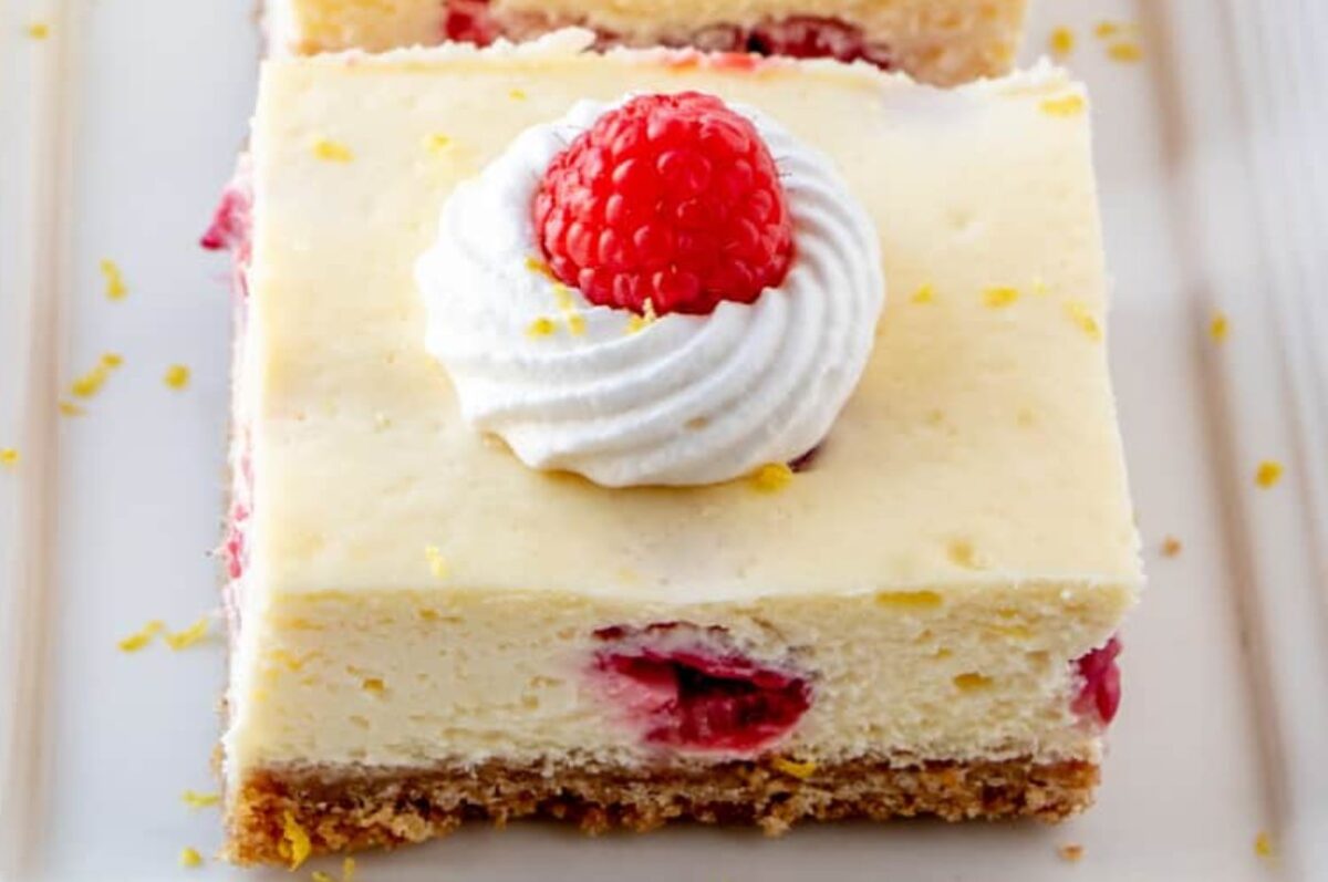 A square cheesecake slice topped with a swirl of whipped cream and a raspberry, featuring a graham cracker crust and visible raspberry pieces within the cream cheese layer, perfect for fans of lemon desserts looking for a fruity twist.
