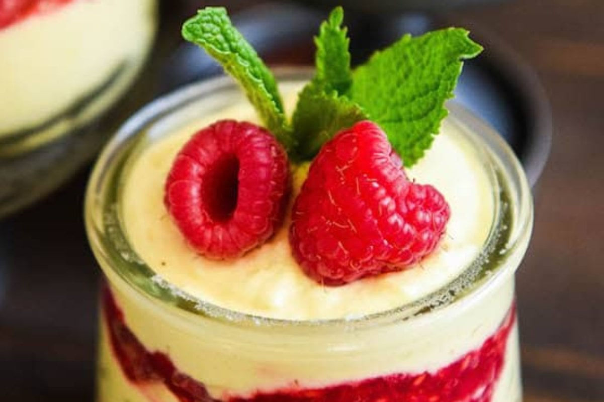 A glass jar filled with a delightful layered dessert, showcasing creamy yellow custard reminiscent of the finest lemon desserts, a vibrant red raspberry layer, and garnished with fresh raspberries and a mint leaf on top.