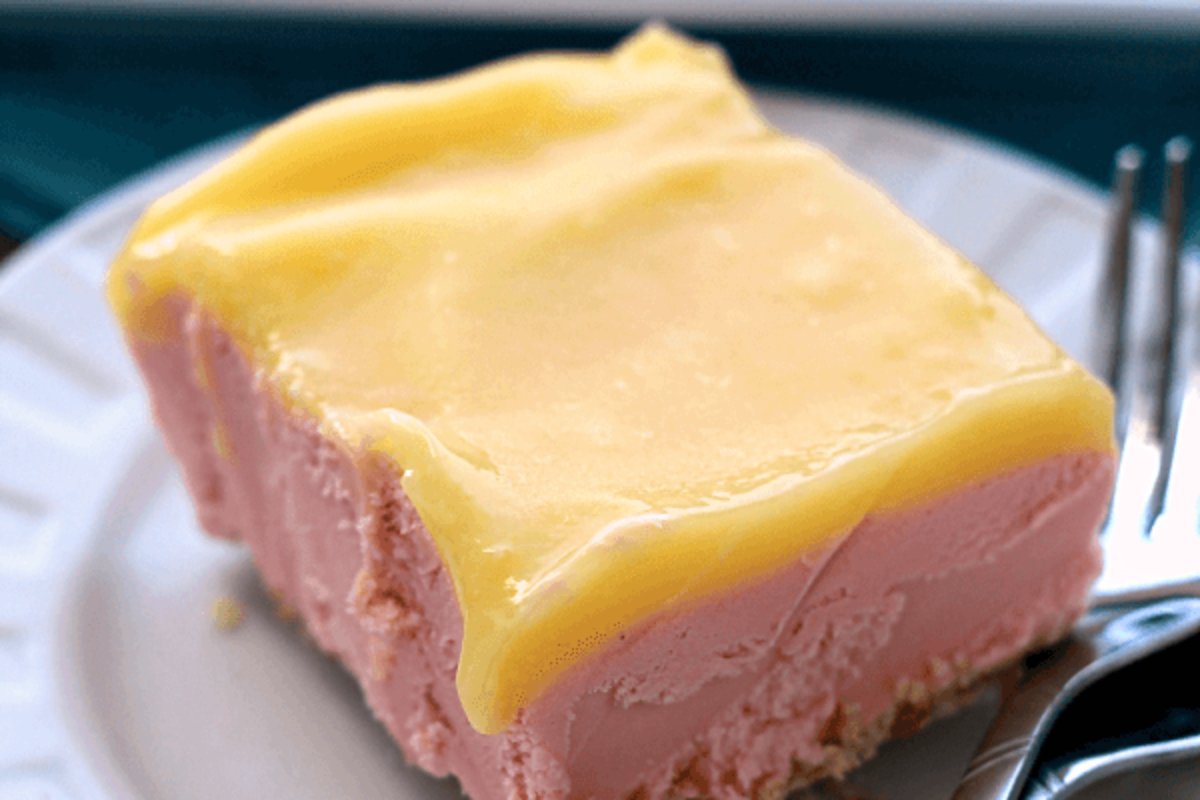 A close-up of a delectable lemon dessert square with a vibrant yellow topping, served on a white plate with a fork and spoon beside it.