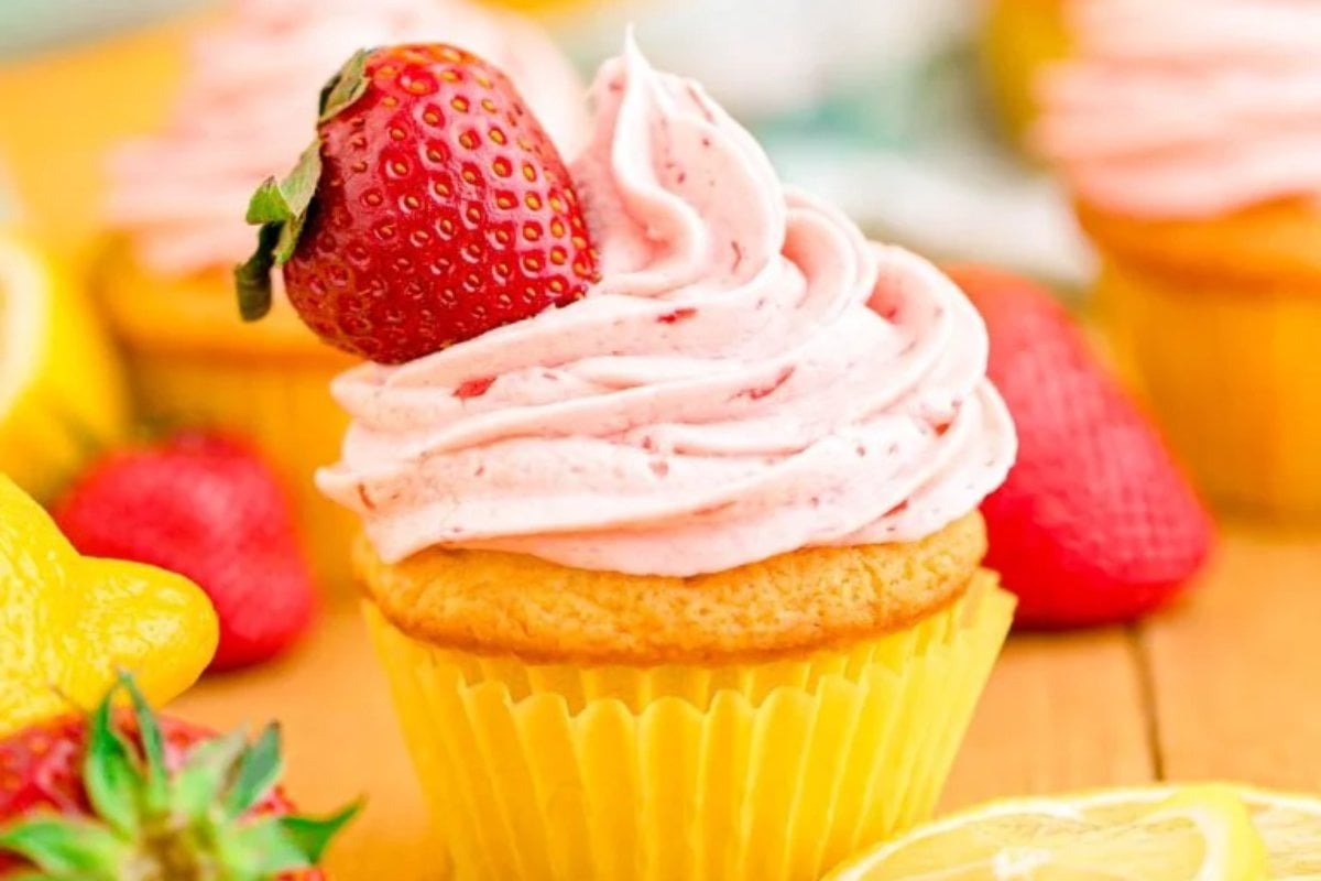 A vanilla cupcake with pink strawberry frosting topped with a whole strawberry, placed in a yellow cupcake liner. Lemon slices and additional lemon desserts and cupcakes are visible in the background.