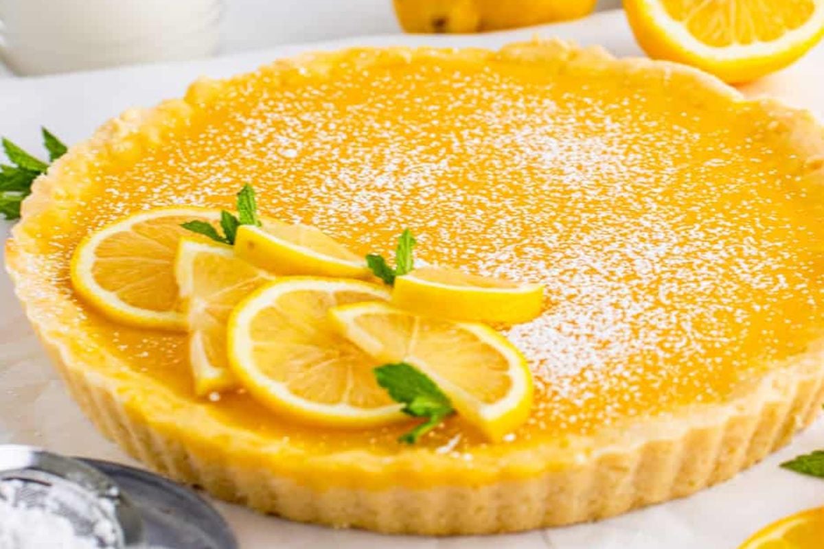 A lemon tart, a perfect example of lemon desserts, garnished with powdered sugar, lemon slices, and mint leaves sits on a white surface.