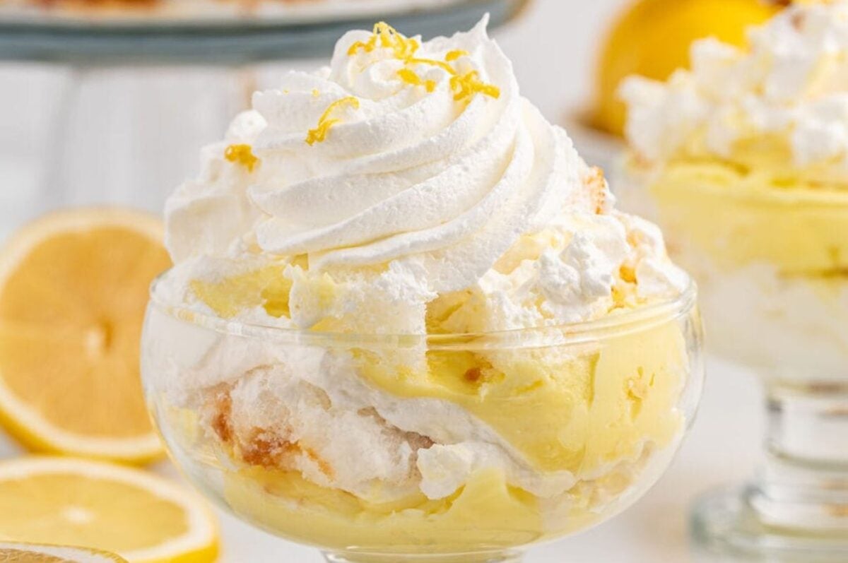 A glass bowl filled with layers of lemon pudding, whipped cream, and topped with lemon zest, epitomizes the allure of lemon desserts. An additional serving sits enticingly in the background with whole lemons nearby.
