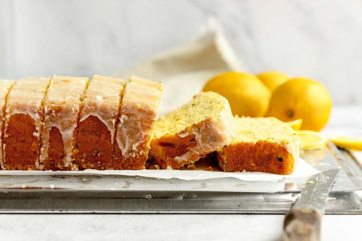 A sliced lemon cake with glaze is placed on a cutting board. Whole lemons are in the background, while a knife rests next to the delightful lemon dessert.