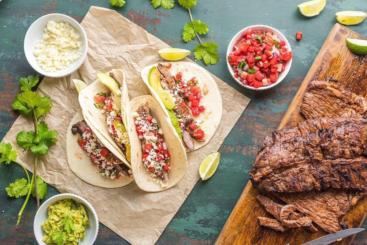 Three beef tacos with diced tomatoes, cheese, and avocado rest on parchment paper, surrounded by bowls of guacamole, pico de gallo, cheese, lime wedges, and grilled meat on a wooden board—perfect for those looking to try delicious steak recipes.