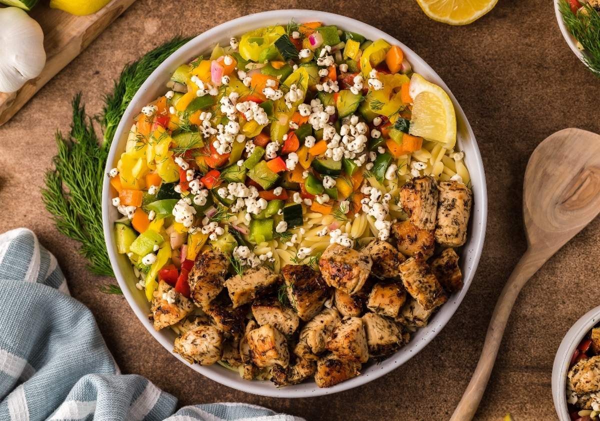 A bowl of colorful salad featuring diced red, green, and yellow bell peppers, cucumbers, and a sprinkle of crumbled cheese, topped with grilled chicken cubes. A lemon wedge is placed on the side, and a wooden spoon lies nearby on a brown surface.