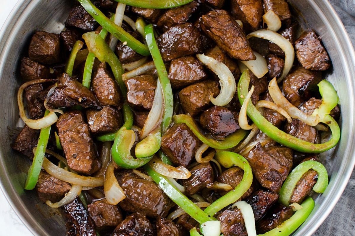 A dish of sautéed beef cubes with green bell peppers and onions in a stainless steel pan, perfect for those exploring steak recipes. The beef appears browned and glazed, while the vegetables are lightly cooked.