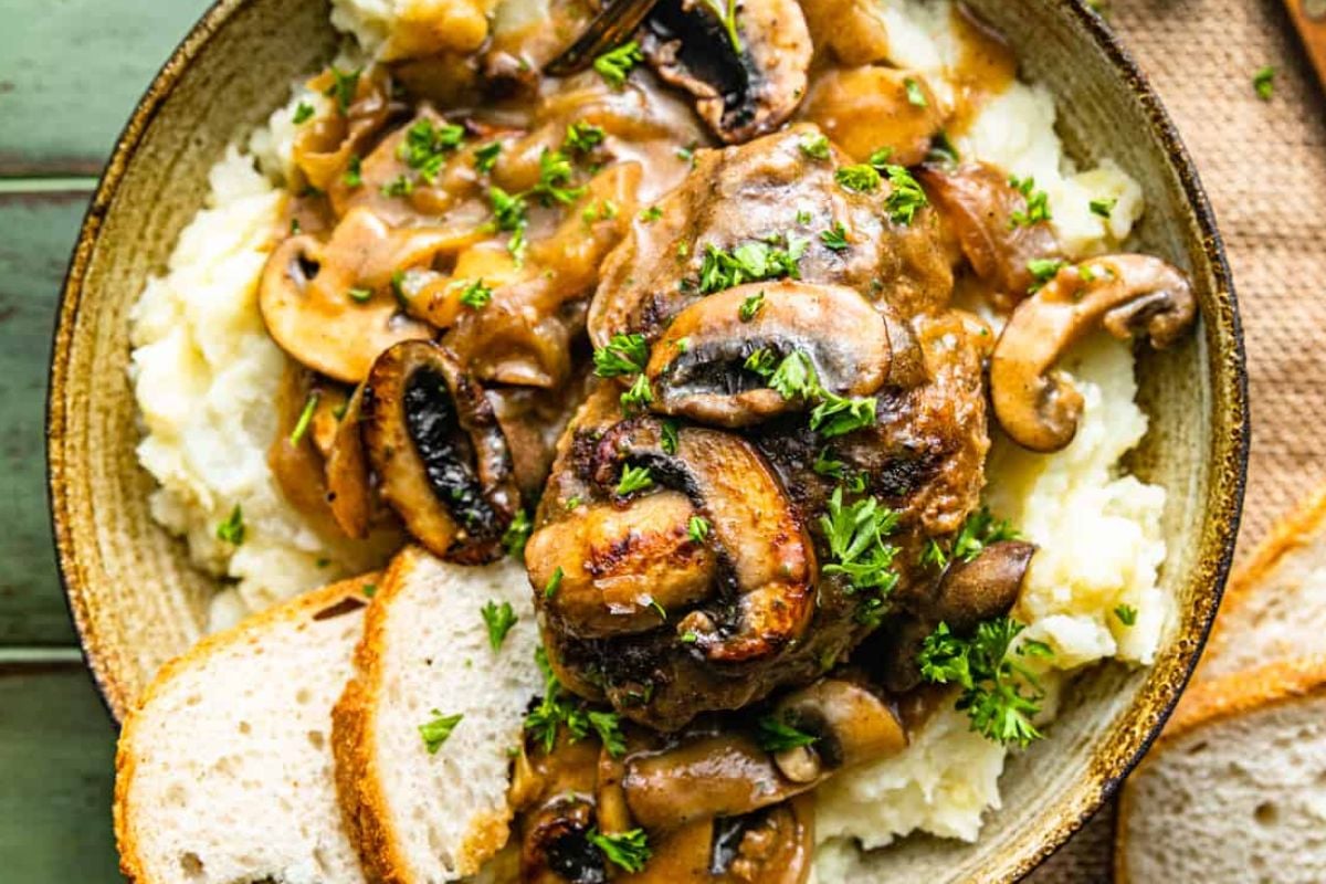 A dish of mashed potatoes topped with a savory meat patty, sautéed mushrooms, and rich gravy, garnished with chopped parsley, served with slices of bread—an ideal addition to your collection of classic steak recipes.