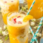 Three glasses of peach Italian cream soda with striped straws, garnished with fruit pieces, and surrounded by flowers and raspberries on a tray.