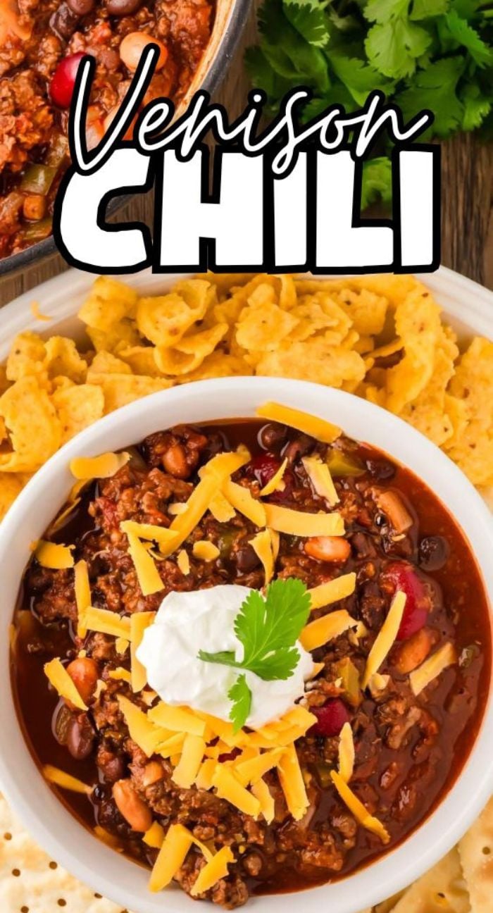 Bowl of venison chili topped with cheese and sour cream, surrounded by corn chips and fresh parsley for the perfect hearty meal.