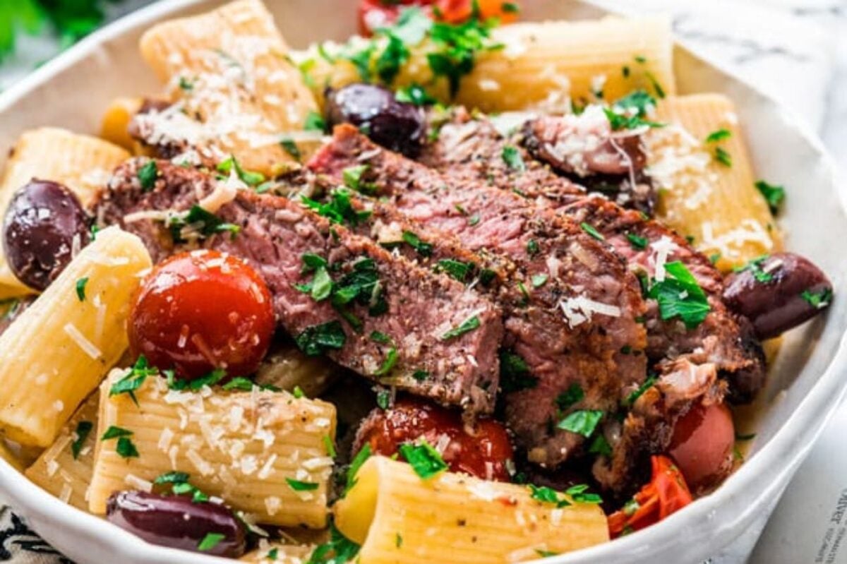 A bowl of rigatoni pasta topped with sliced grilled steak, cherry tomatoes, kalamata olives, and fresh chopped parsley, sprinkled with grated cheese—a delightful addition to your favorite steak recipes.