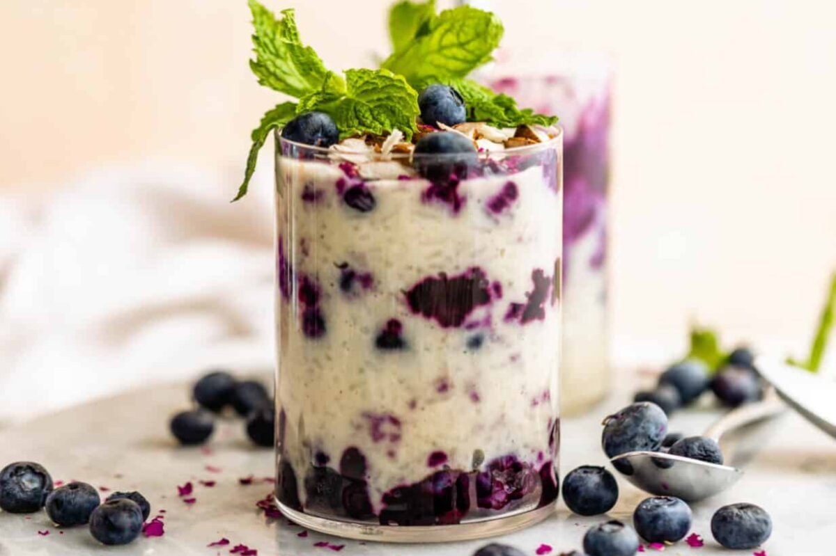 A glass of layered blueberry chia pudding, a delightful treat from our collection of blueberry recipes, is topped with fresh blueberries and mint leaves. Scattered blueberries and a spoon rest artfully on the pristine white surface.