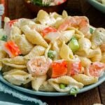A plate filled with a creamy seafood pasta salad, featuring shell-shaped pasta, shrimp, crab meat, peas, and celery cubes. A light green napkin is partially visible to the left.
