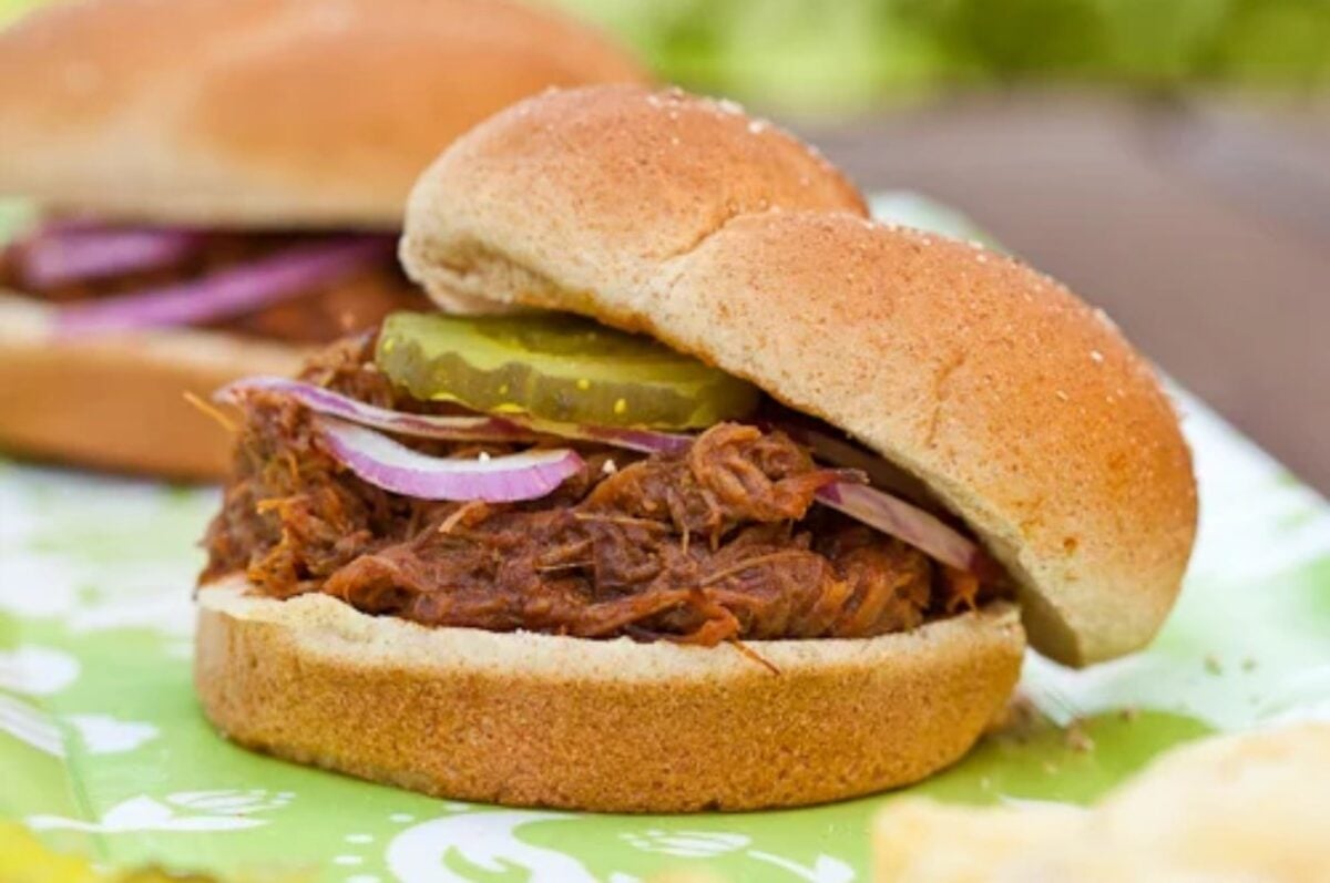 A slow cooker dinner delight: a pulled pork sandwich with pickle slices and red onion on a bun, placed on a green and white surface. Another sandwich is blurred in the background.