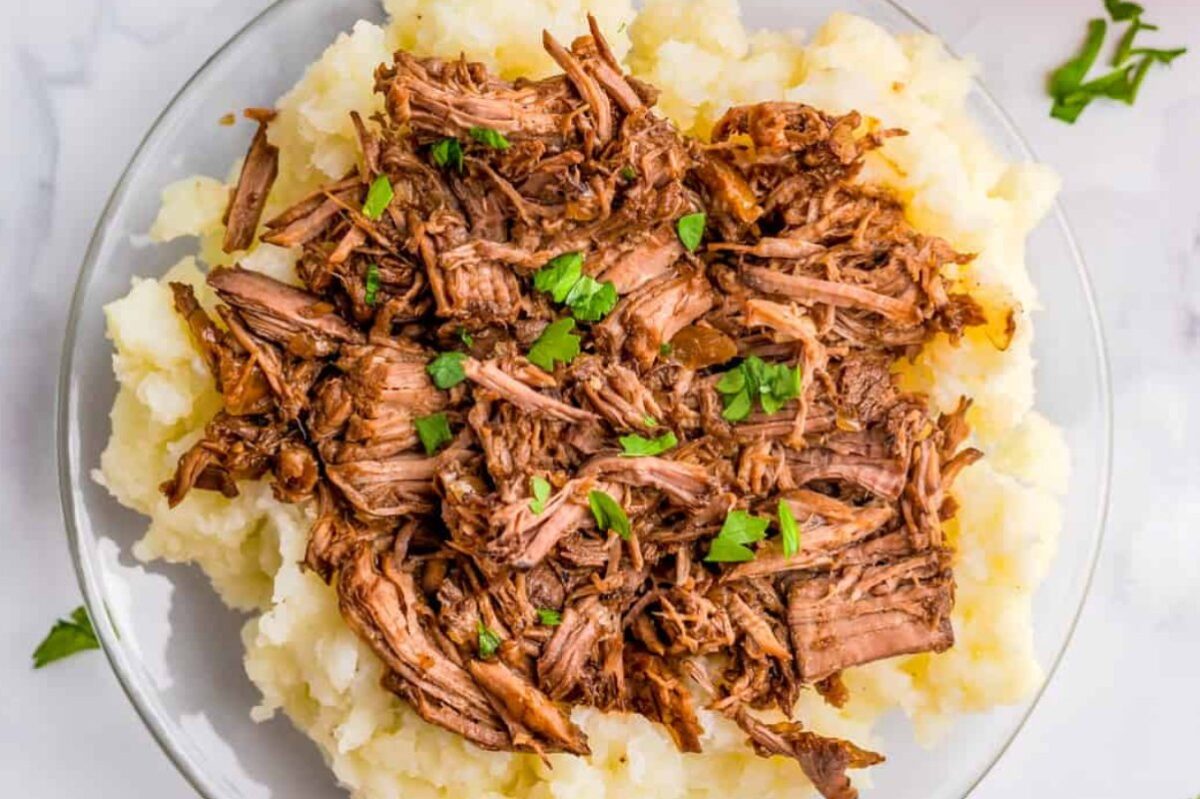 A plate of mashed potatoes topped with slow-cooked shredded beef, garnished with chopped parsley.