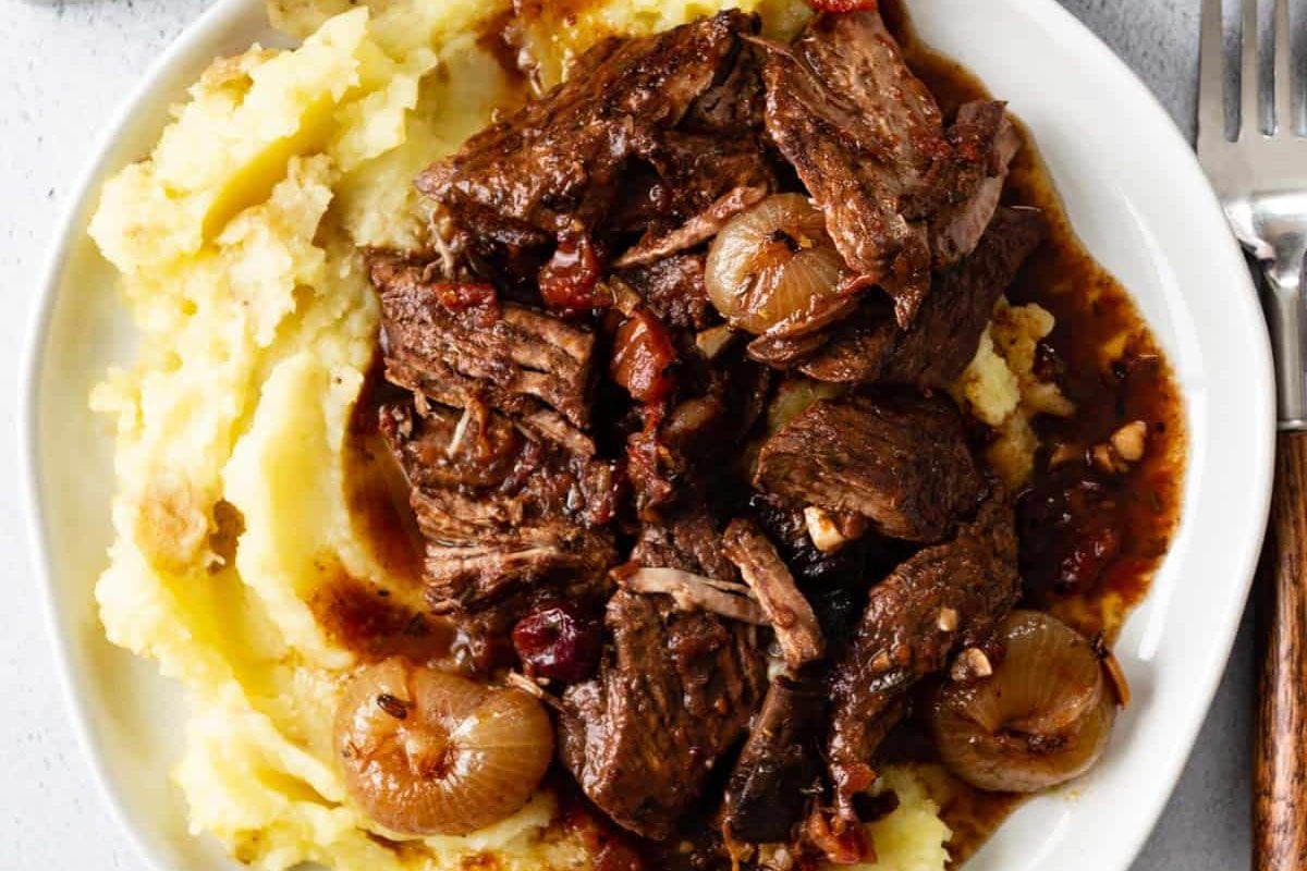 A plate of mashed potatoes topped with braised beef chunks and pearl onions in a dark gravy, reminiscent of hearty slow cooker dinners.
