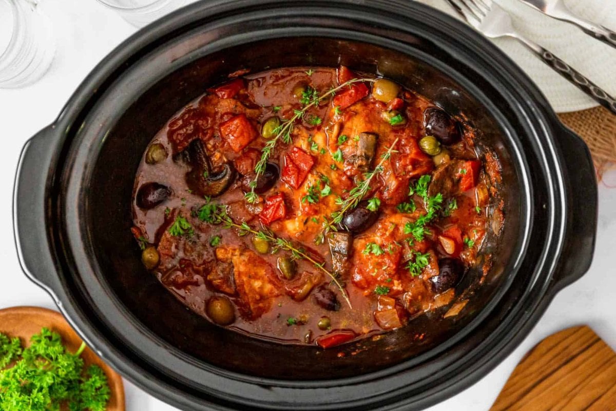 A slow cooker filled with a chunky stew mixture of chicken, tomatoes, olives, and herbs, viewed from above. Embrace the essence of slow cooker dinners with a small dish of fresh herbs beside it on the left.