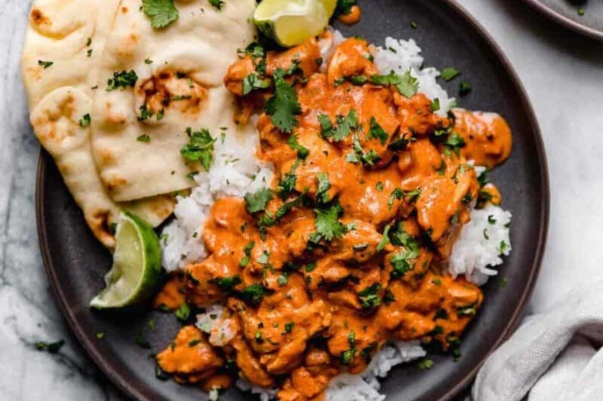 A plate featuring rice topped with creamy orange curry, garnished with cilantro and lime wedges, alongside a piece of naan bread - a perfect slow cooker dinner.