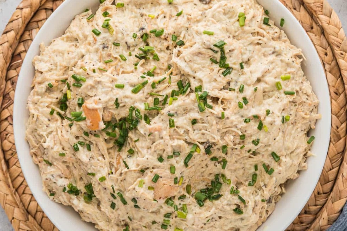 A bowl of creamy, shredded chicken dip garnished with chopped chives, perfect for slow cooker dinners, placed on a woven mat.
