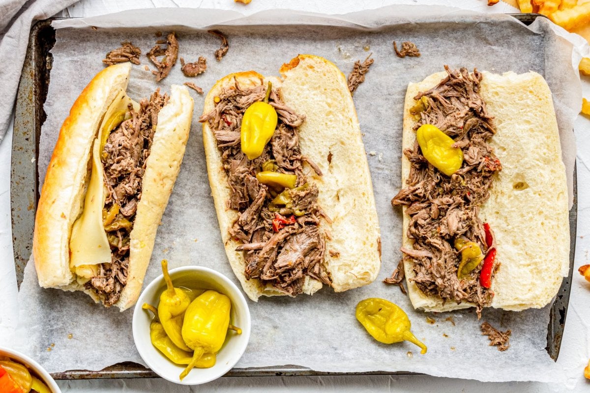 Three Italian beef sandwiches with shredded beef and pepperoncini on a lined baking tray next to a small bowl of pepperoncini&mdash;perfect examples of delicious slow cooker dinners.