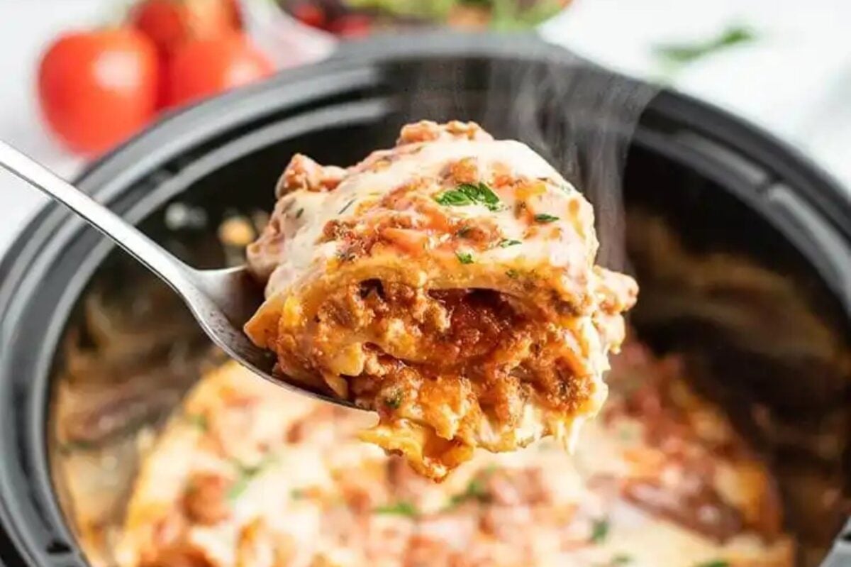 A close-up of a fork lifting a portion of cheesy, layered lasagna from a slow cooker. Steam rises from the freshly cooked dish, embodying the essence of comforting slow cooker dinners. In the background, two tomatoes lend a splash of color to this delicious scene.