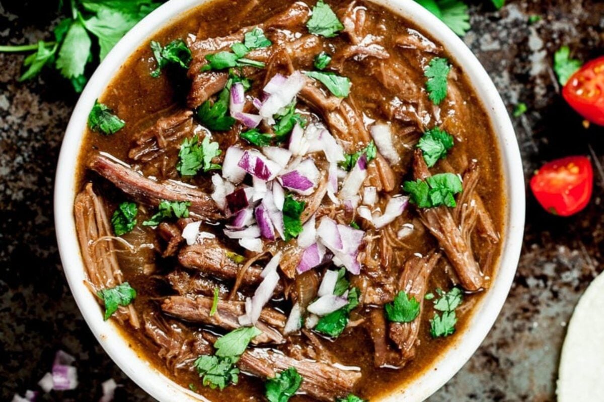 A close-up of a bowl of beef stew, a comforting favorite among slow cooker dinners, garnished with chopped red onions and cilantro, with halved cherry tomatoes in the background.