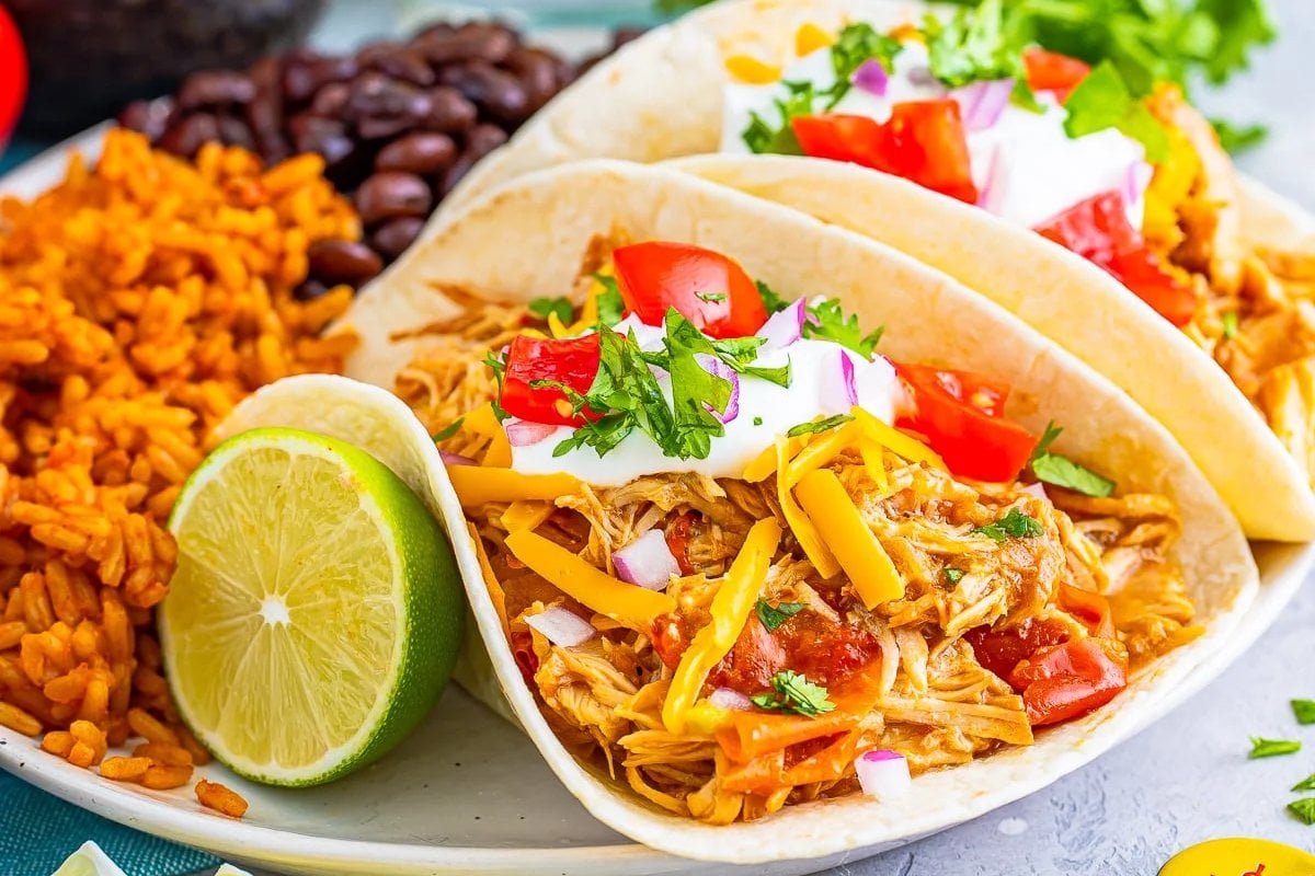 Plate with two chicken tacos topped with cheese, tomatoes, onions, cilantro, and sour cream, accompanied by a lime wedge, Mexican rice, and black beans&mdash;a perfect addition to your lineup of slow cooker dinners.