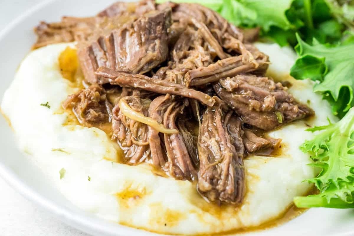 Plate of tender, slow-cooked shredded beef served over creamy mashed potatoes with a side of leafy greens.