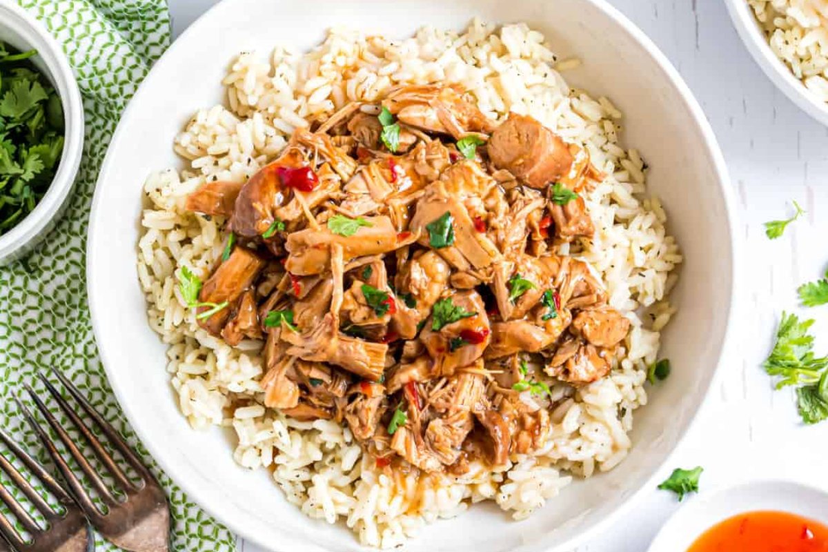 A white bowl filled with white rice, topped with shredded, sauce-covered meat from one of those delicious slow cooker dinners, garnished with chopped herbs. A fork, a small bowl of herbs, and a green cloth napkin are nearby.