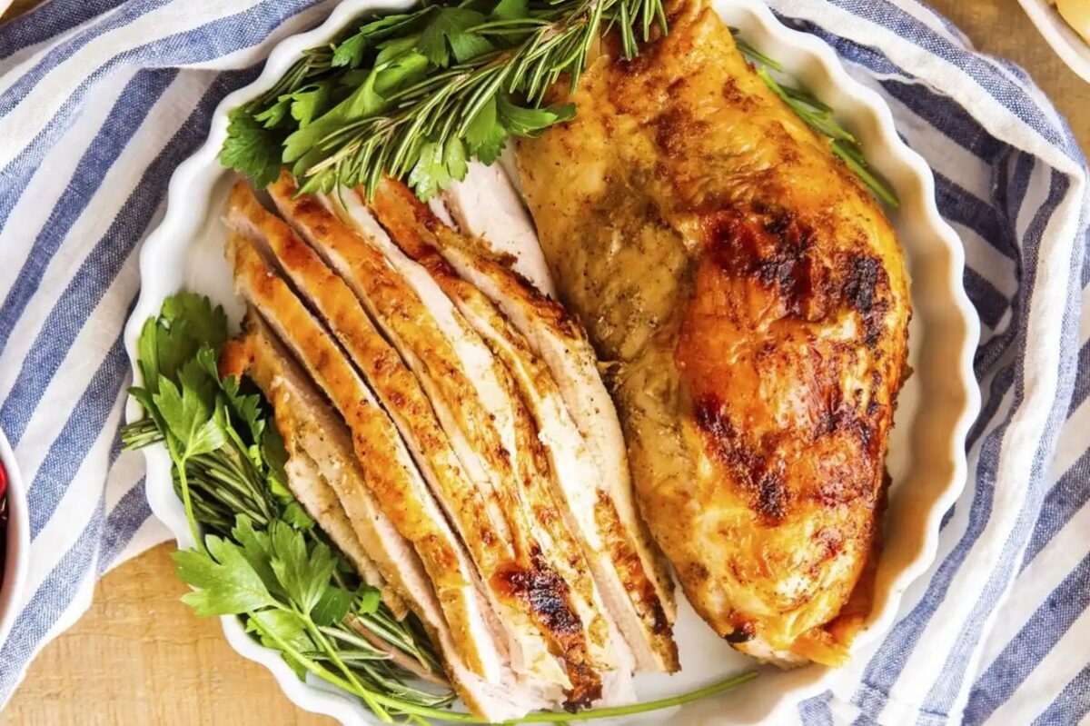 A white dish with a slow-cooked turkey breast, half sliced and half whole, garnished with fresh rosemary and parsley, placed on a blue and white striped cloth.