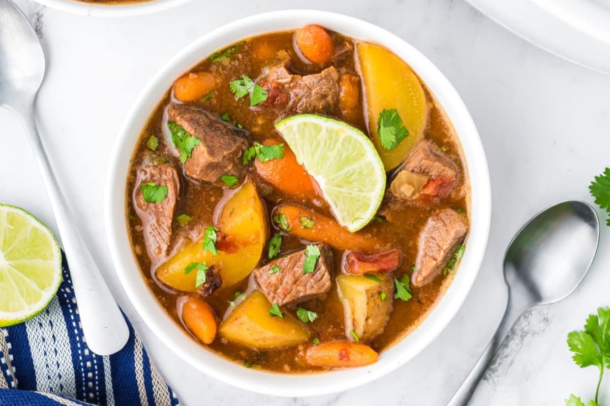 A bowl of beef stew with potatoes, carrots, and topped with a lime wedge and chopped parsley. A spoon is next to the bowl on a white surface.