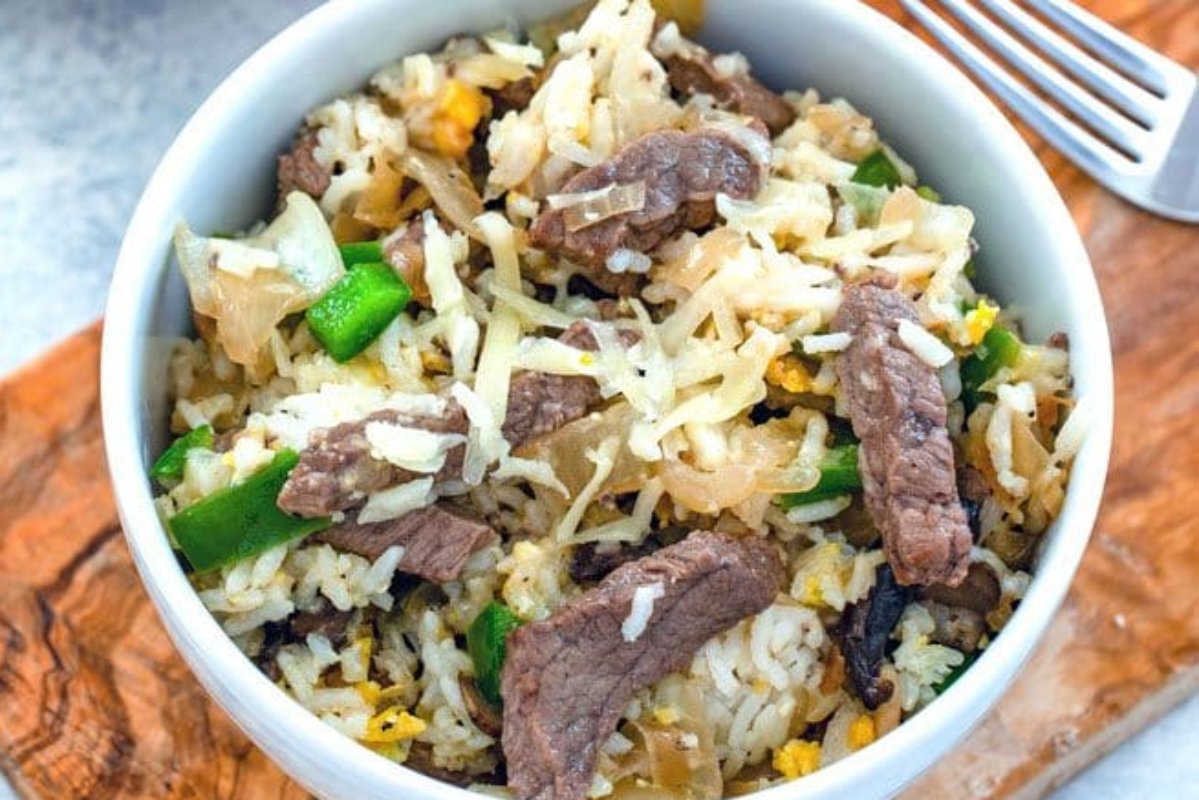 A bowl of stir-fried rice with strips of beef, green bell peppers, shredded vegetables, and bits of scrambled egg on a wooden surface next to a fork—perfect for those looking to diversify their steak recipes.