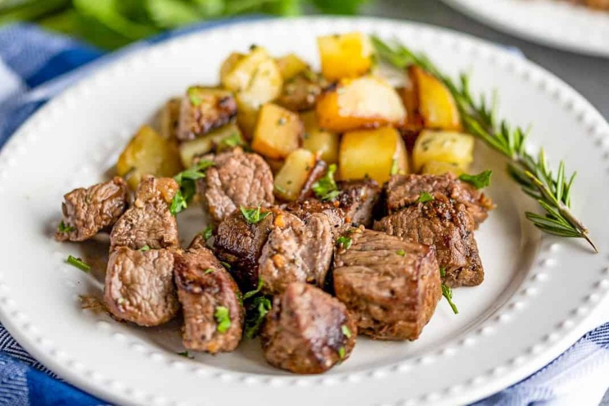 A white plate with seasoned beef cubes and roasted diced potatoes garnished with herbs, reminiscent of classic steak recipes, accompanied by a sprig of rosemary.