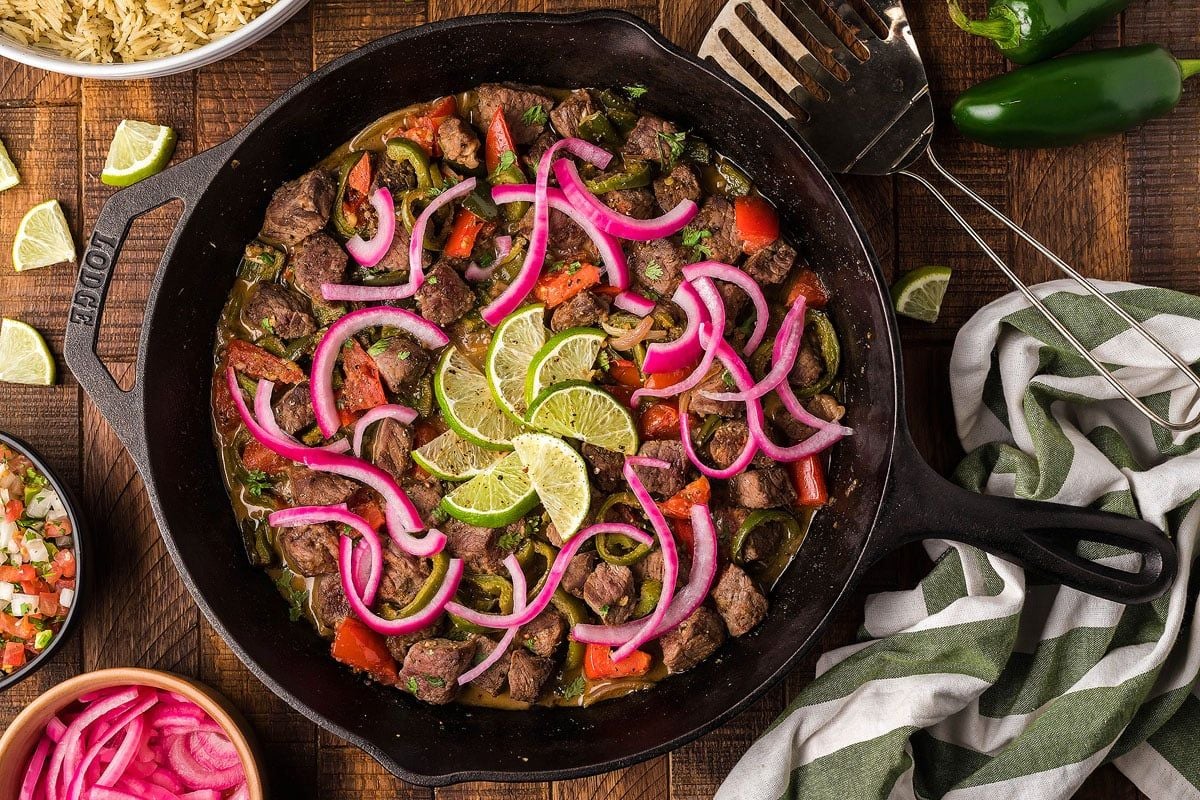 A cast-iron skillet filled with beef fajitas, garnished with lime slices and red onions. Surrounding the skillet are additional toppings, rice, a kitchen towel, and a spatula—essential elements for perfect steak recipes.